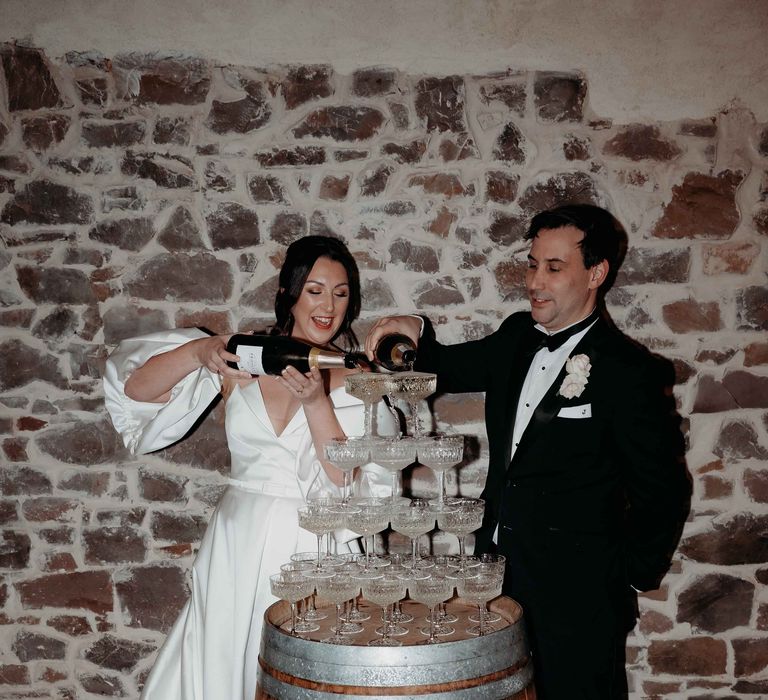 Bride in white silk wedding dress pours champange over champagne tower with groom in black tuxedo