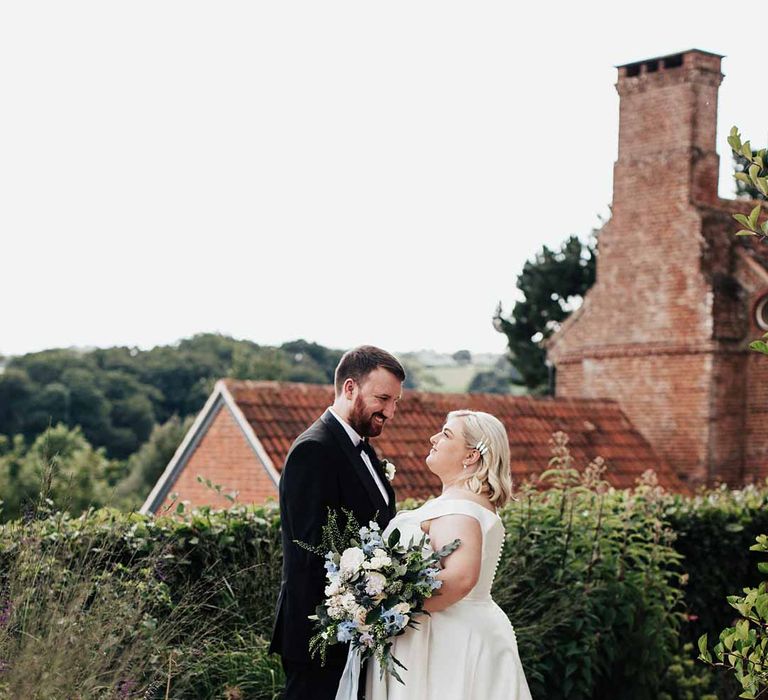 Groom in classic black grooms three piece tuxedo with white rose boutonniere and black bowtie standing with bride in satin off the shoulder wedding dress holding white and blue eucalyptus, foliage, garden rose, gardenia, light blue delphinium and dried flower bridal bouquet tied with dusky blue ribbon 