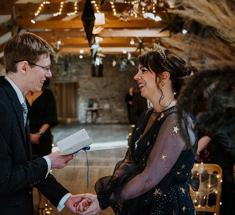 Groom in navy suit laughs and holds hands with the bride as he reads out his personal wedding vows from a DIY vow booklet 