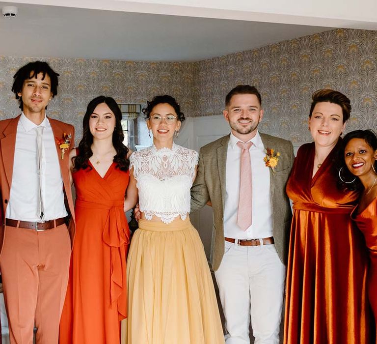 Bride wearing white mesh lace bridal crop top and yellow wedding skirt standing with bridesmaids in mismatched bridesmaid dresses and wedding guests in yellow and orange wedding theme