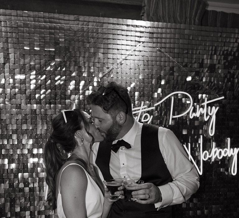 Bride and groom share a kiss with there cocktails in front of neon wedding sign and glitter wall 