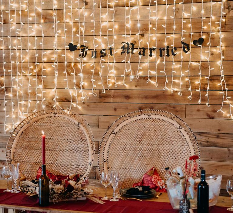 Wedding high table with wicker chairs by gothic wedding tablescape with deep red table runner, pampas grass and dried flower table centrepieces, red tapered candles in black bottles, fairy light wall and black just married sign