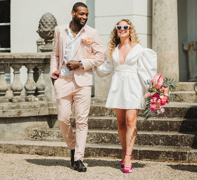 Groom walking in pastel pink wedding suit with orchid buttonhole with the bride in a short wedding dress with puff statement sleeves 