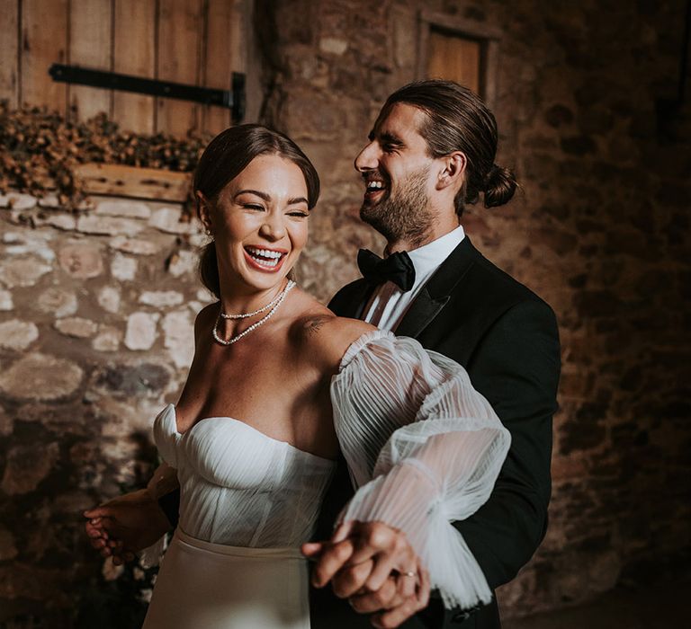 Bride in corset style Made With Love wedding dress with detachable sleeves posing with the groom in a black tuxedo 