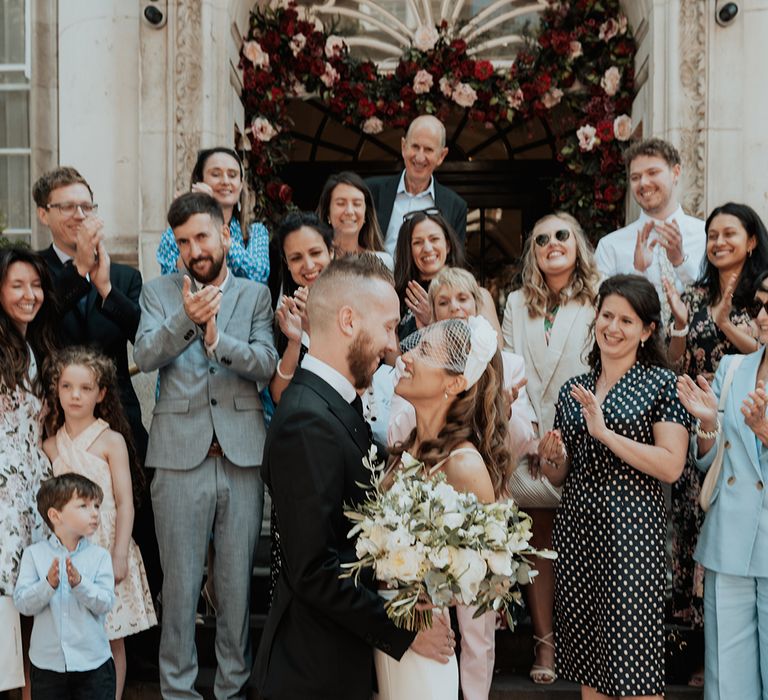 bride in a birdcage veil smiling at her groom in a black suit on the steps of Chelsea Town hall 