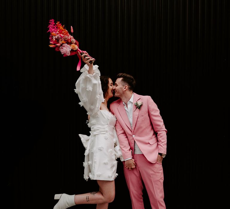 The bride holds her hot pink dried wedding flower bouquet in the air as she kisses the groom in a pink suit 