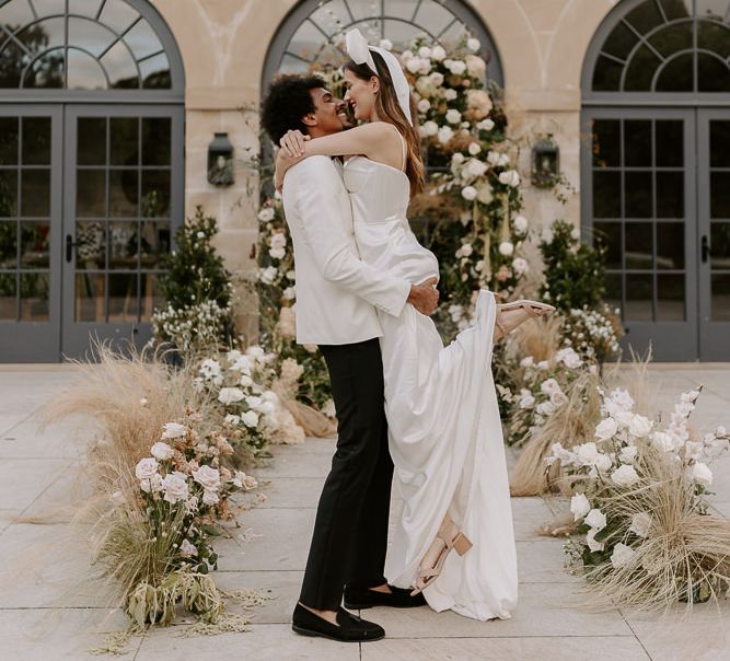 Black groom in a white tuxedo jacket picking up his bride in a satin wedding dress and bow headdress