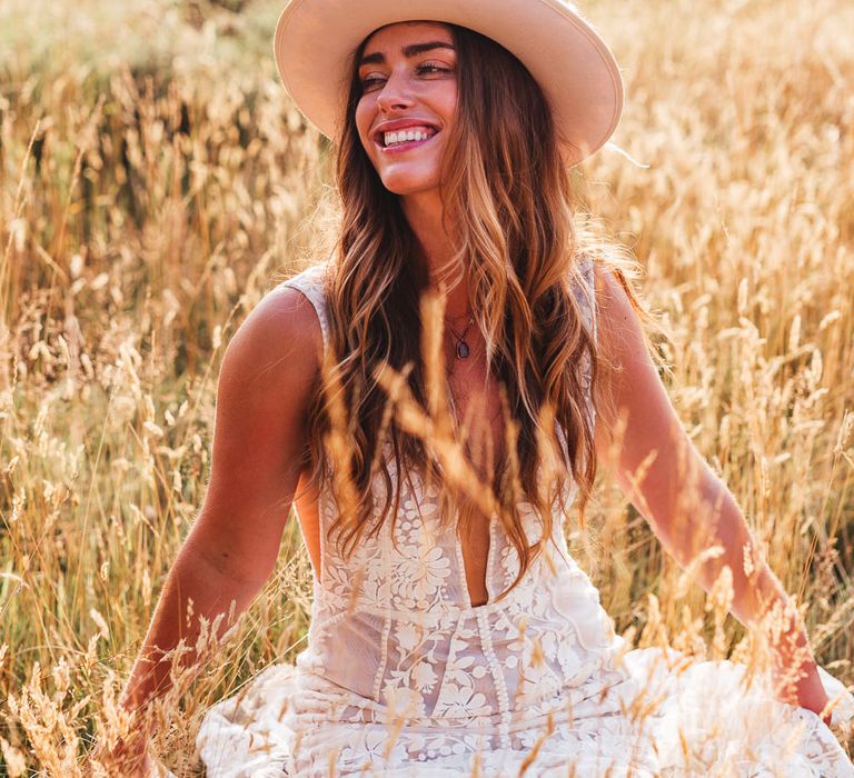 Bride sits in fields during golden hour wearing white fedora