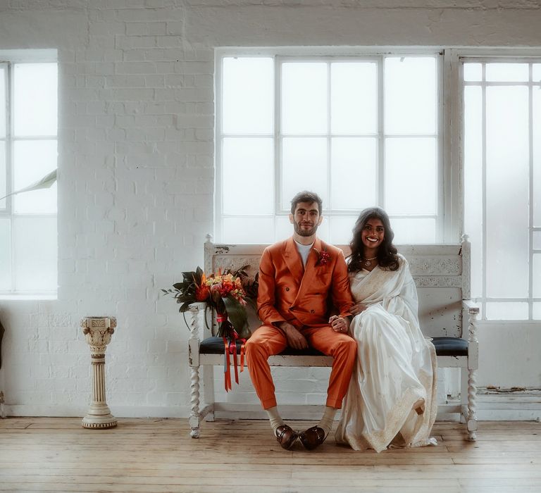 Bride in a white sari and groom in an orange suit sitting on a chair at their industrial wedding 