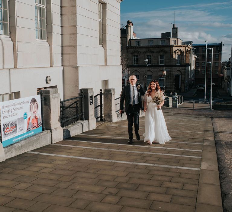 Father walks bride in white wedding dress holding dried flower bouquet to town hall