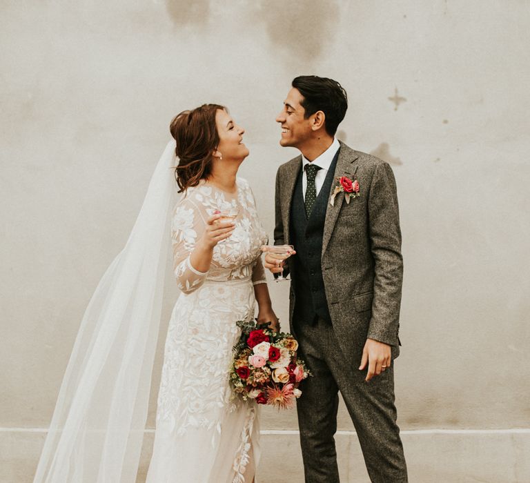 Wedding portrait of the bride and groom smiling at each other in a lace wedding dress with long sleeves and front split and grey wool suit 