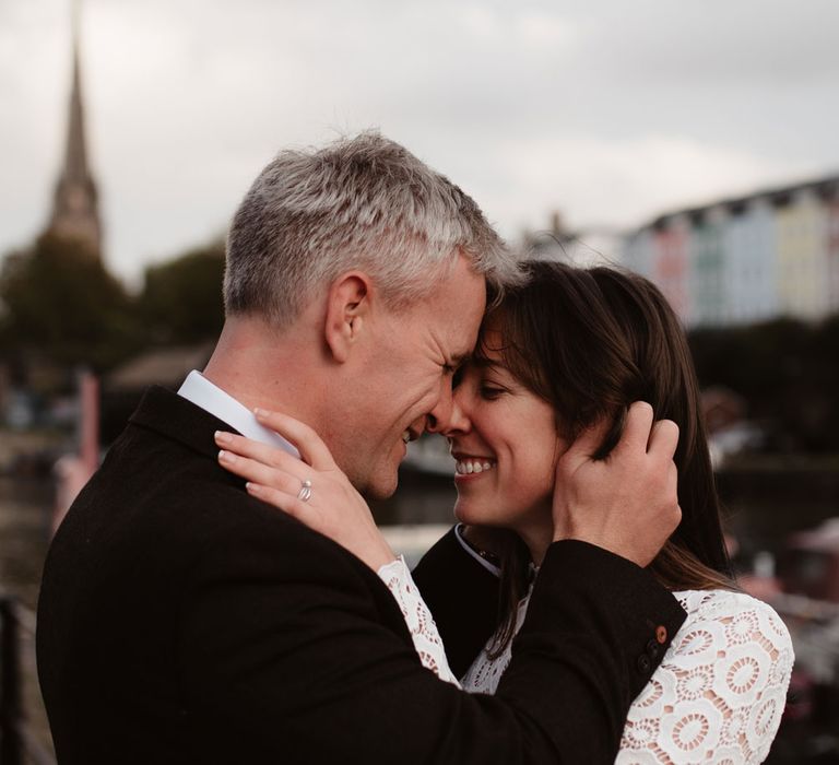 Smiling groom in brown woollen blazer and smiling bride in long sleeved lace top Self Portrait wedding dress and silver wedding ring embrace by Bristol Harbourside