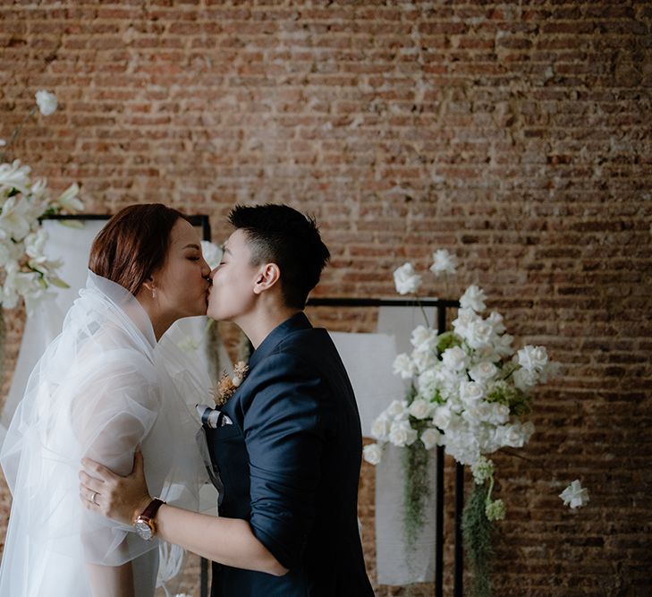 Asian Lesbian wedding ceremony with bride in a navy suit kissing her bride in a white minimalist dress and veil