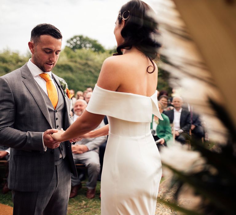 Bride in white strapless Vagabond wedding dress holds hands with groom in grey Hugo Boss suit with waistcoat and yellow tie stand holding hands at the top of the aisle