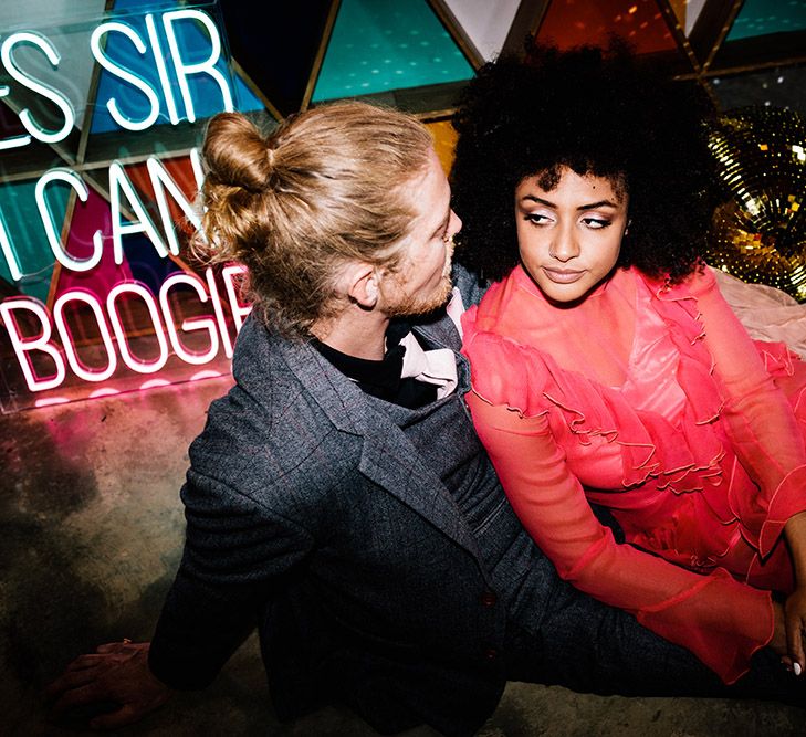 Stylish bride and groom sitting on the dance floor with a neon wedding sign and disco balls 