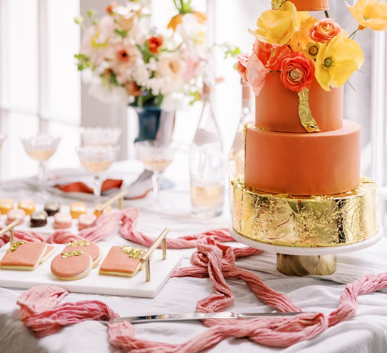 Four tier wedding cake with varying colours of orange frosting, complete with floral decor and gold leaf