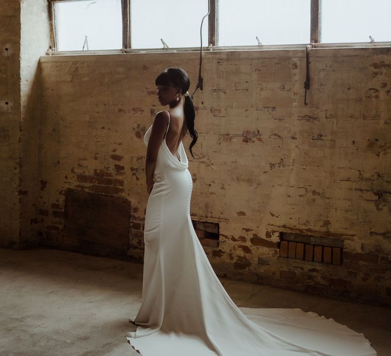 A Black bride in a white low back wedding dress stands with her back to the camera. The train is long and she wears her long hair in a lo ponytail that cascades down her back.