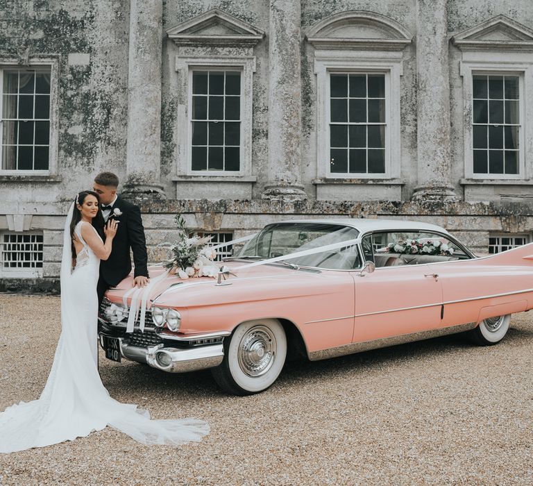 Bride in white Pronovias wedding dress and veil wraps arms around groom in black suit whilst leaning back on pink Cadillac in front of Came House Dorset after their wedding