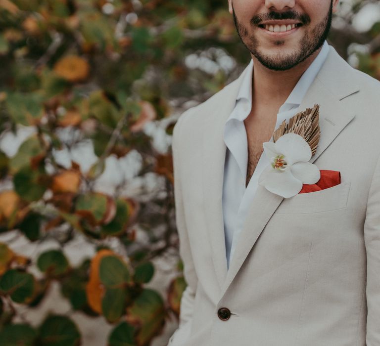 Groom smiles as he wears light coloured suit with open shirt, red pocket square and orchid buttonhole 