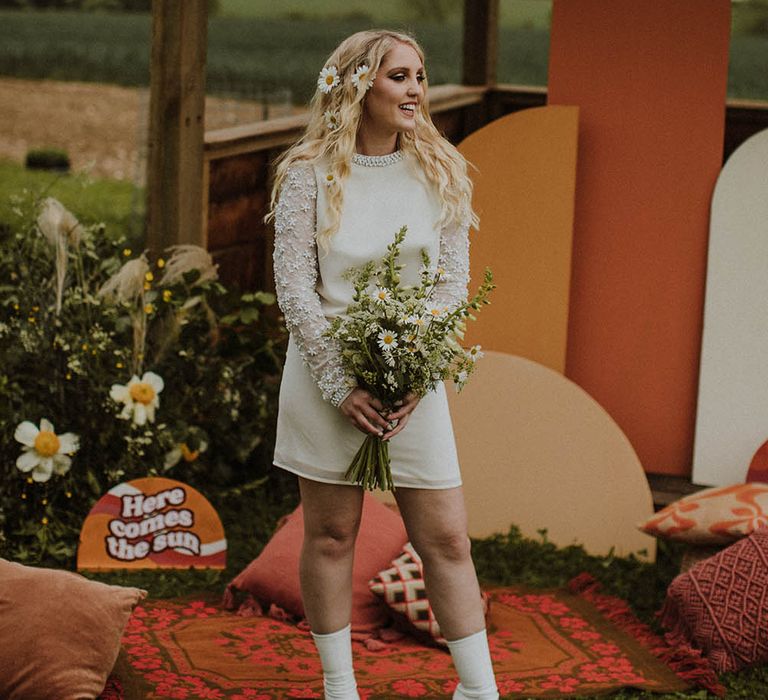 Beautiful boho bride in a short wedding dress with long applique sleeves, white boots and flowers in her hair holding a giant daisy bouquet