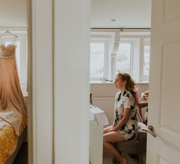 Bride sits in bedroom getting ready for barn wedding ceremony whilst pearl wedding dress hangs in the room next door