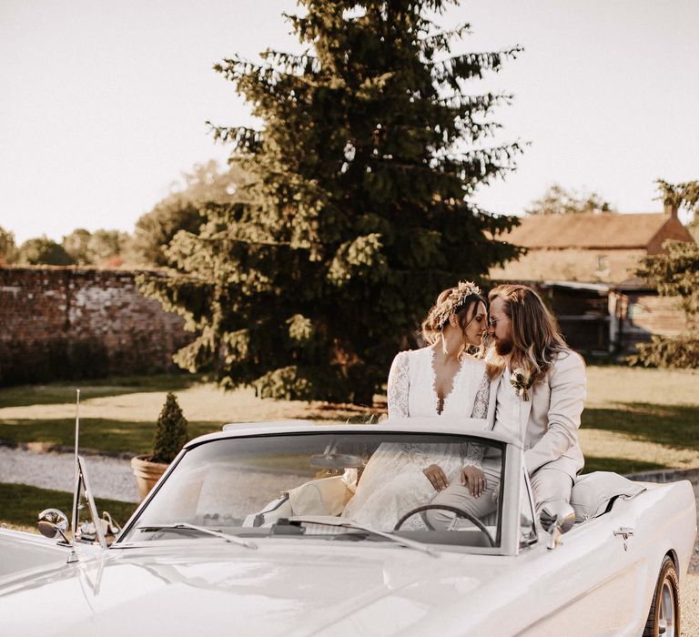 Boho bride and groom in a lace wedding dress and beige suit sitting on the back of their white mustang wedding car 