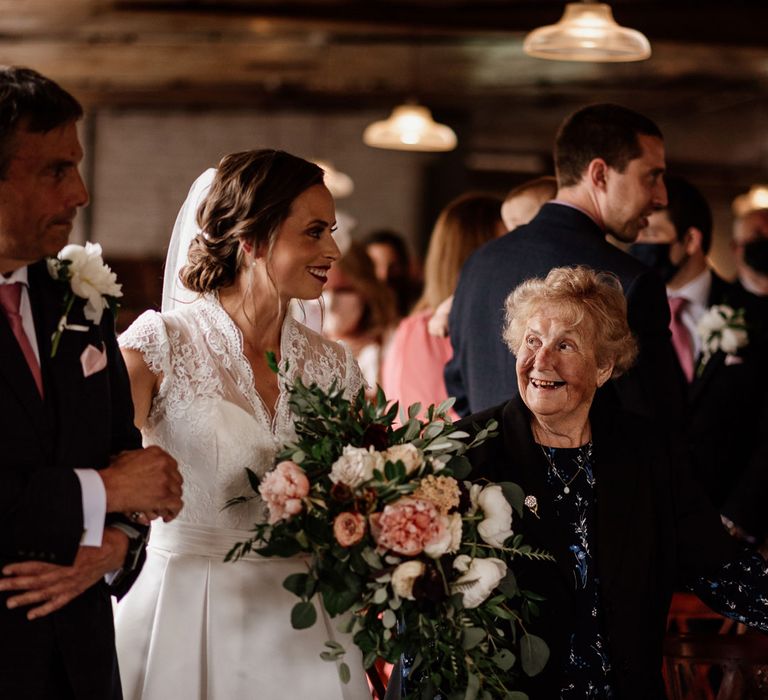 Bride in white lace cap sleeve wedding dress and veil holds pink white and green bridal bouquet as she walks down the aisle arm in arm with man in checked suit and pink tie at The West Mill Derby