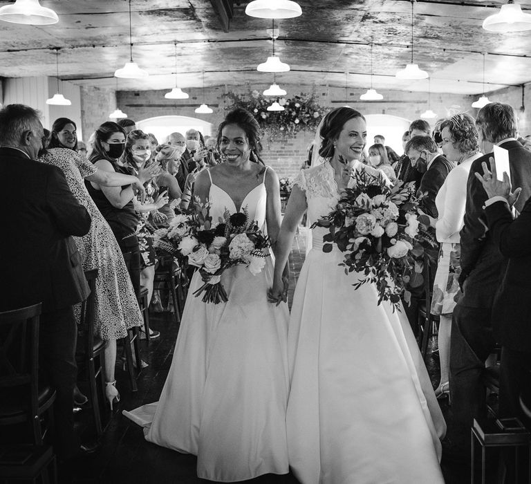 Smiling bride in white cami wedding dress holds hands with bride in white lace capped sleeve wedding dress as they both carry white, pink and green bridal bouquets down the aisle at The West Mill Derby