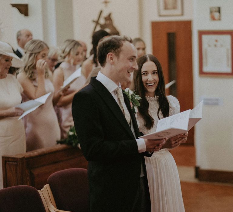 Laughing bride in white lace puffed sleeve Daalarna wedding dress looks at groom in black morning coat as they hold hymn books during church ceremony