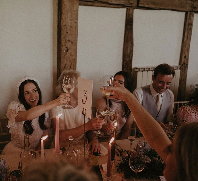 Bride in white lace puffed sleeve Daalarna wedding dress and bridal headband raises a glass of white wine to wedding guests as they sit at long table for wedding breakfast at Loseley Park wedding in Surrey