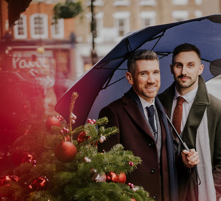 Grooms stand with one another underneath umbrella on their wedding day in London as it rains