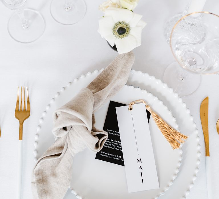 Two white plates are stacked and rest on a white table cloth. There is a neutral coloured tied napkin that rests next to two pieces of overlapping card that is tied with a gold tassel. The top card reads Matt'. The cutlery is white and gold and the glassware has gold rims. There is an anemone in a bud vase above the plates.