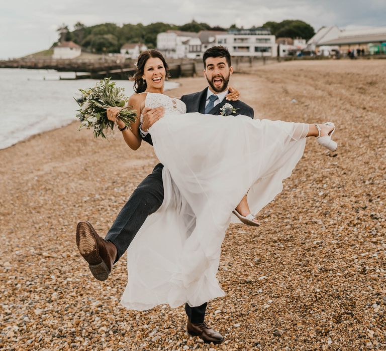 Groom lifts bride along beach front across the sand 