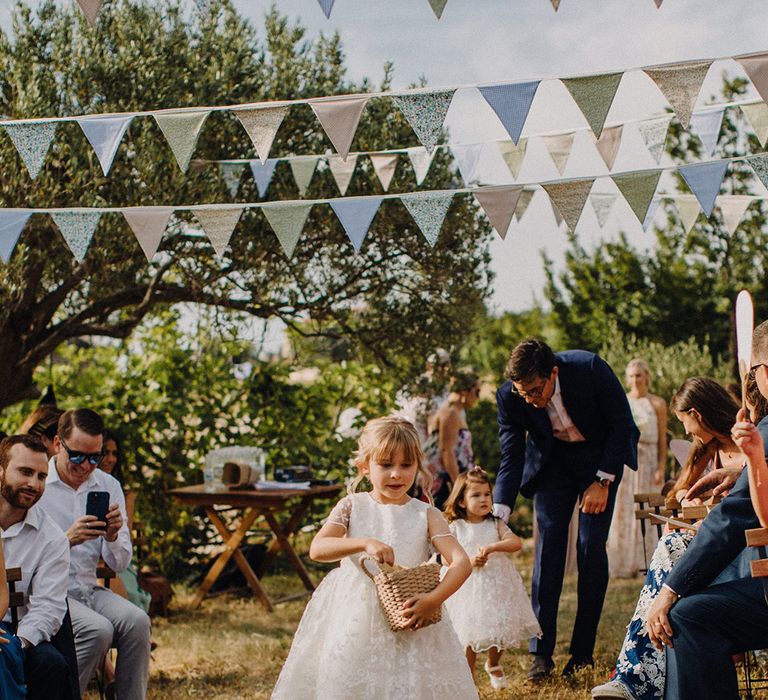 Flower girls walk down the aisle as they carry wicker baskets filled with confetti as bunting hangs above them