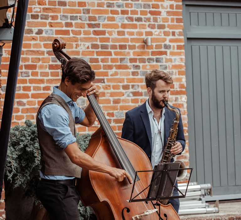 Man in black chinos, blue shirt and brown waistcoat plats double bass next to man in blue blazer jacket and light blue shirt playing saxophone outside at Wasing Park wedding