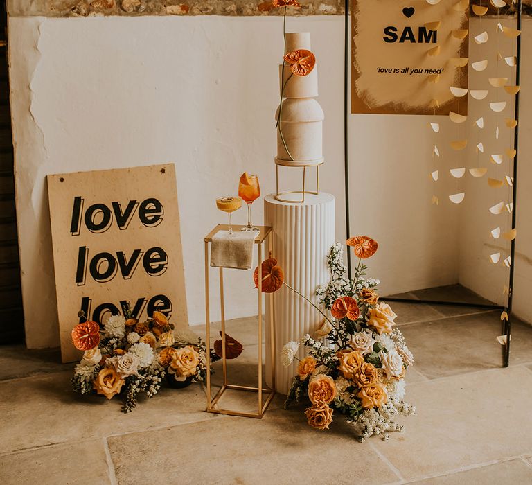 Wedding cake display and decor with tall wedding cake on a white plinth, and retro signs hanging from a copper frame 