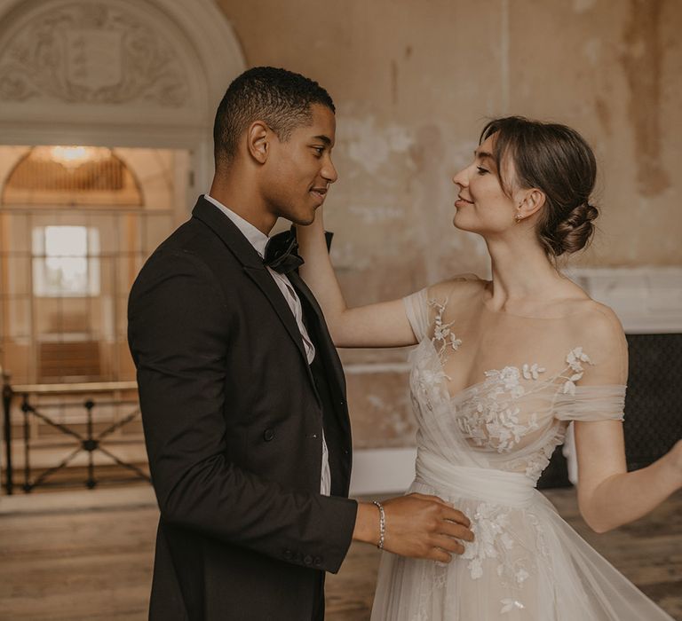 Bride in a tulle wedding dress with illusion neckline and lace detailing touches her grooms face in a black tie suit 