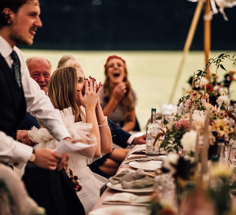 Bride in white shirt, blue waistcoat and patterned tie makes speech during garden marquee wedding reception in Cornwall as bride in Halfpenny London Mayfair dress and wedding guests laugh