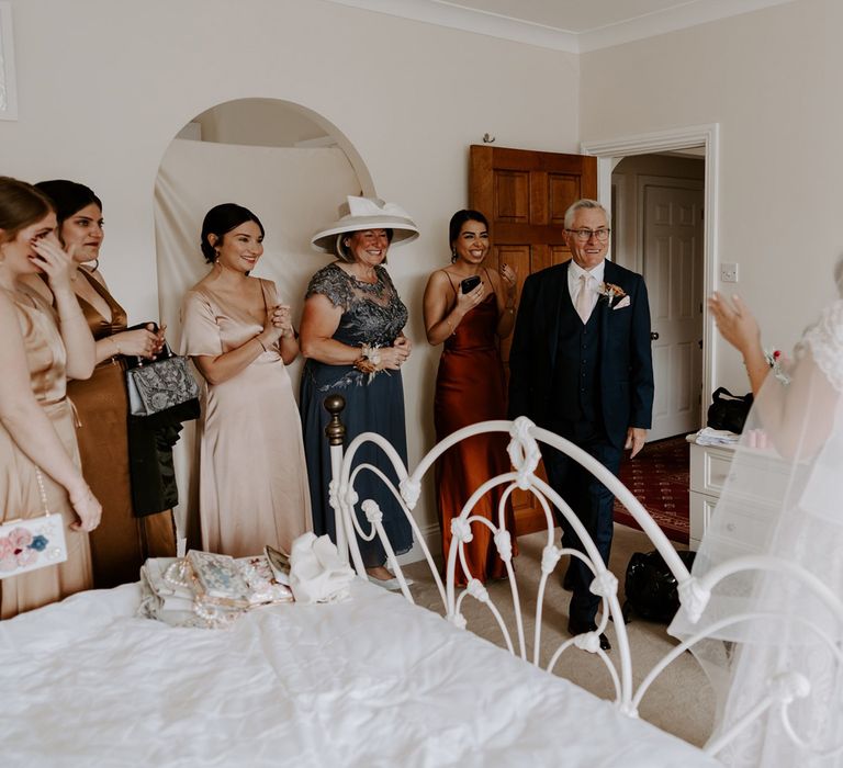 Wedding guests stand in bedroom and smile at bride in homemade lace wedding dress and veil before DIY garden wedding ceremony in Bedfordshire