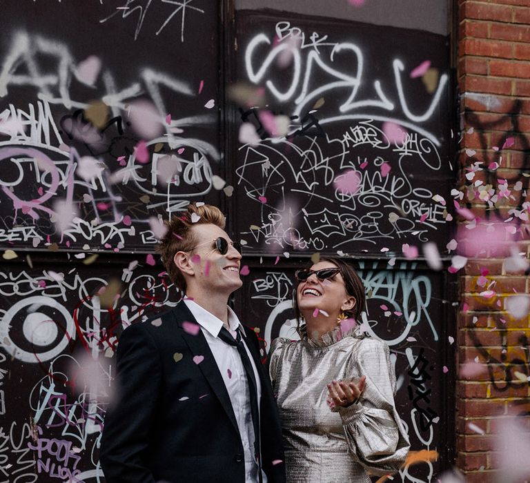 Pink confetti falls onto Bride and Groom as they stand in front of brick wall full of artwork