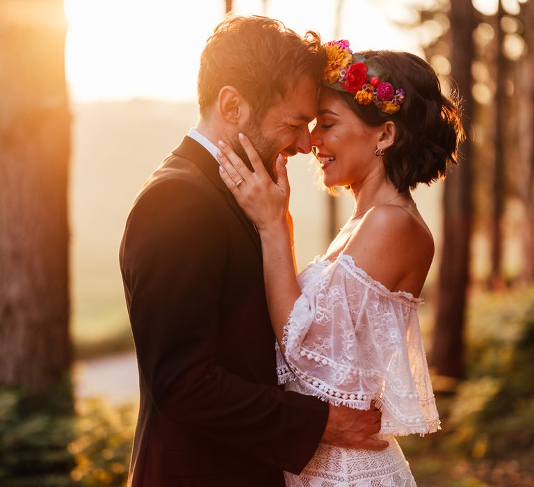 Smiling bride in off the shoulder Grace Loves Lace wedding Dres sand flower crown stands with groom in black suit in woodland during golden hour