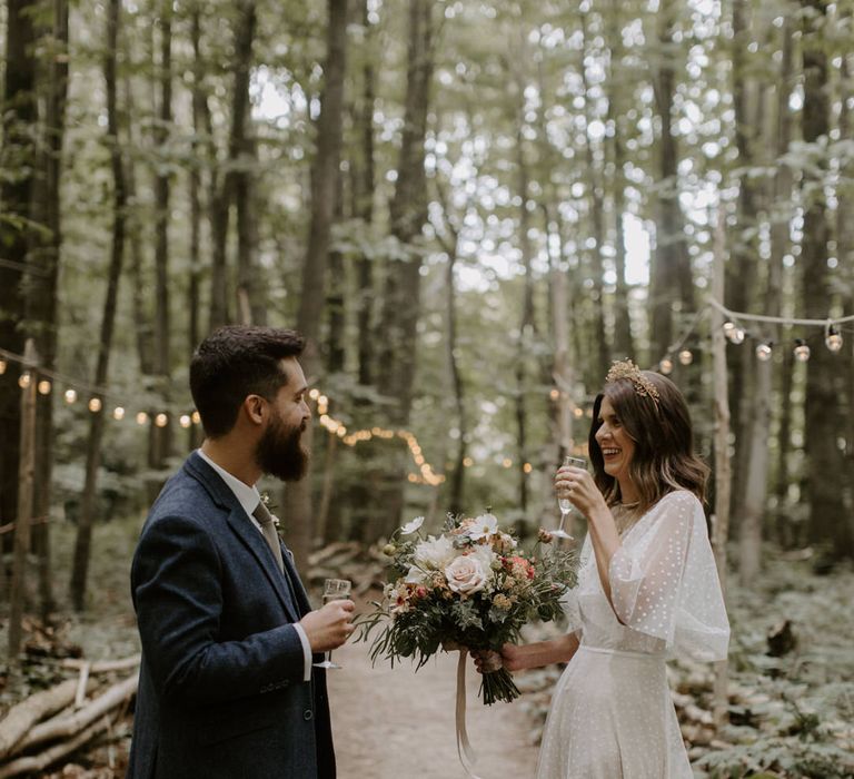 Bride & groom laugh and drink champagne with one another in woodlands at The Dreys wedding venue