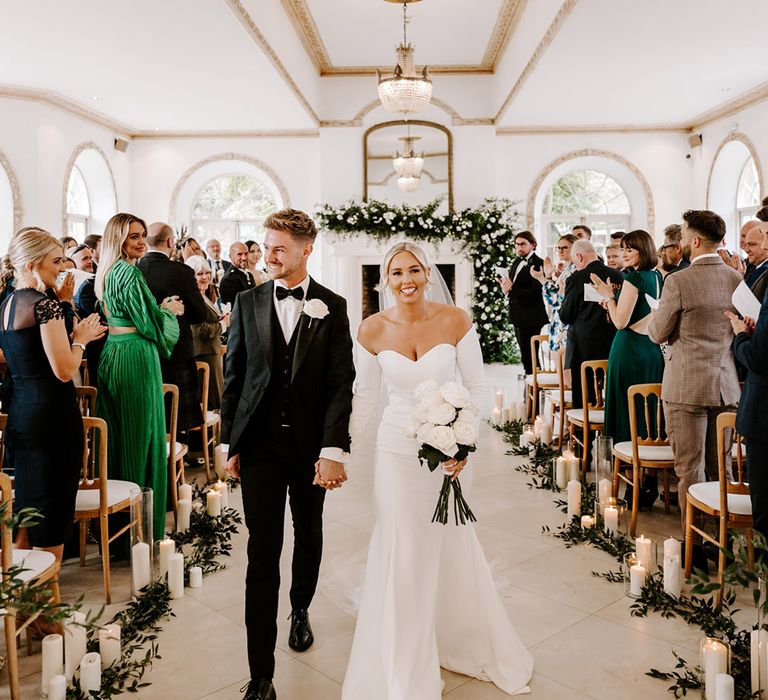 Bride in a fitted wedding dress with detachable long sleeves and groom in a tuxedo descending up the aisle as husband and wife