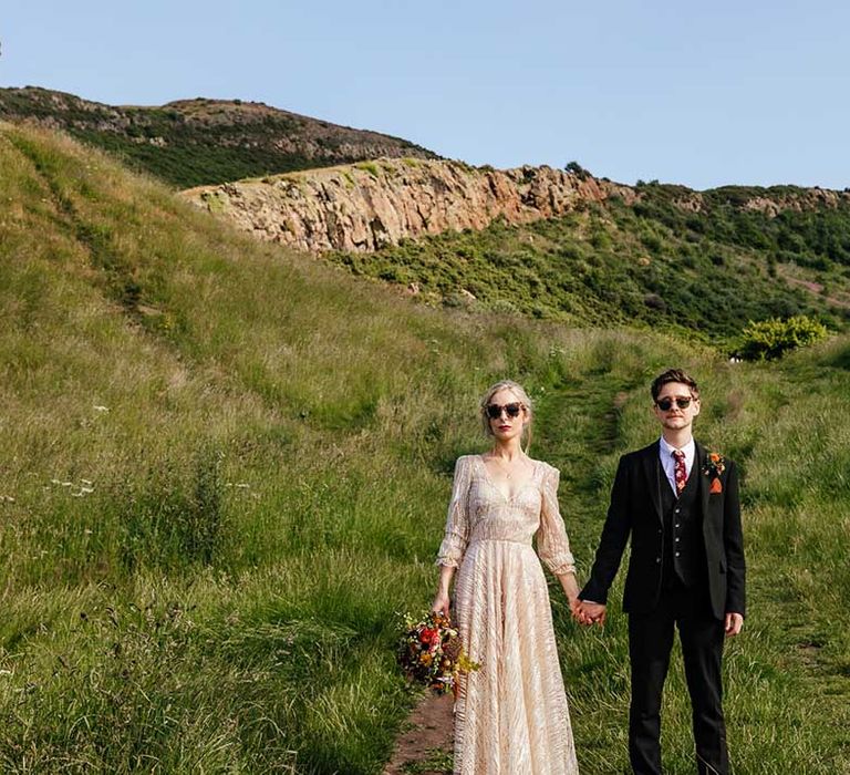 Bride & groom hold hands as they look toward the camera whilst wearing sunglasses 