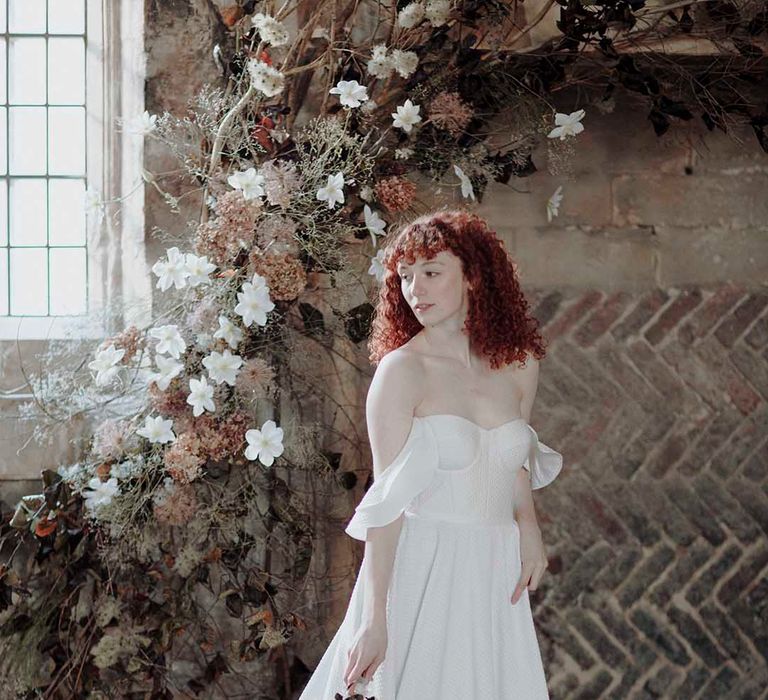 Bride red curly hair in a strapless princess wedding dress holding a minimalist wedding bouquet by a dried flower floral arrangement 