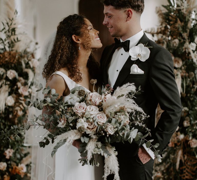 Bride & groom under floral arch whilst bride holds floral bouquet with green foliage