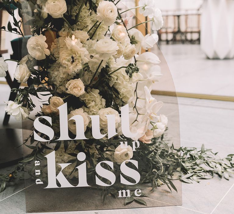 Shut Up and Kiss Me smokey acrylic wedding sign standing in front of a white floral arrangement with hydrangeas, ranunculus and anthuriums. 