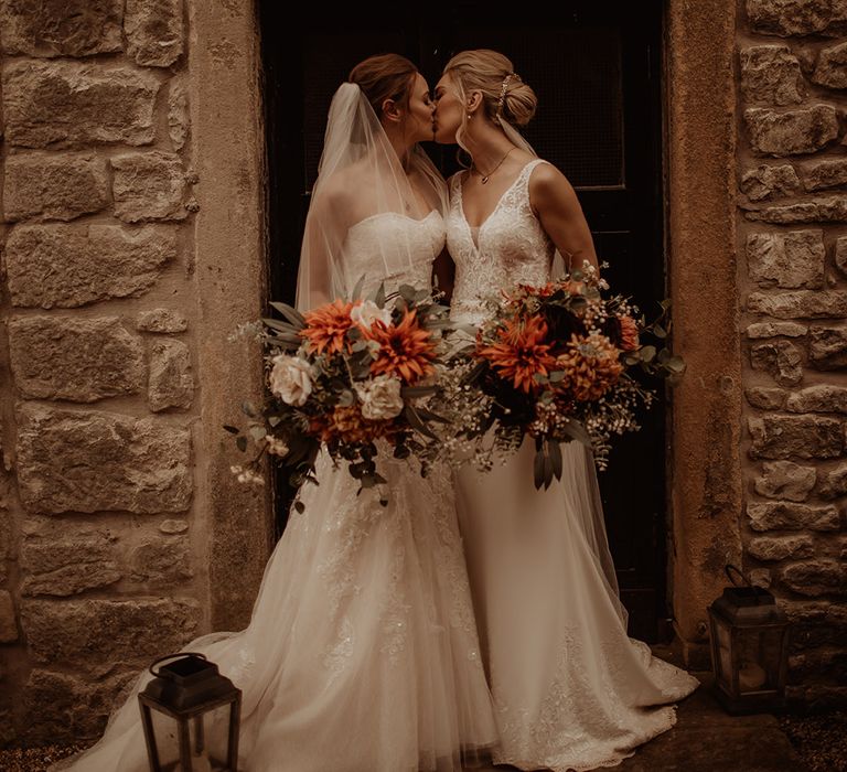 Brides kiss outdoors as they hold floral bouquets