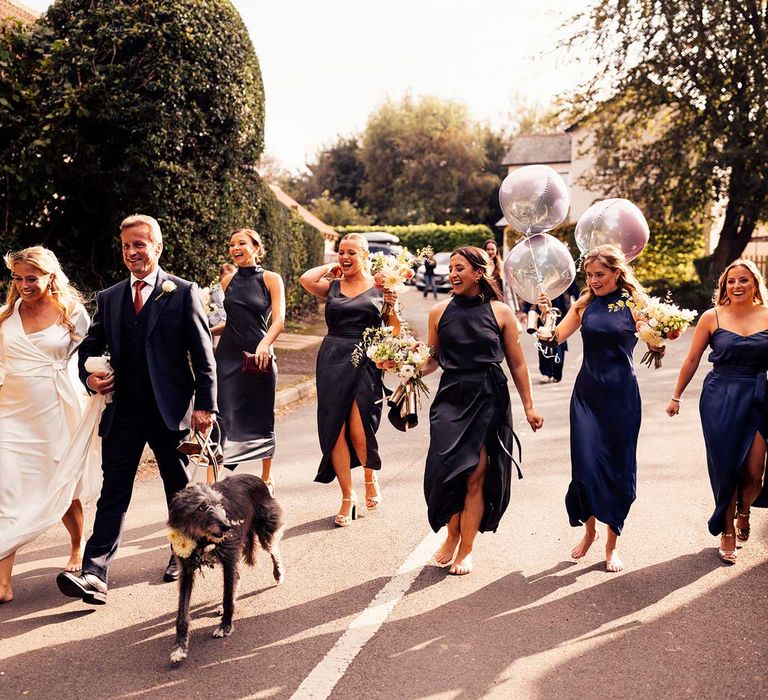 Bride in wedding dress and wrap top carrying bouquet walks barefoot down road with man in blue suit, bridesmaids in navy satin bridesmaid dress and dog towards church