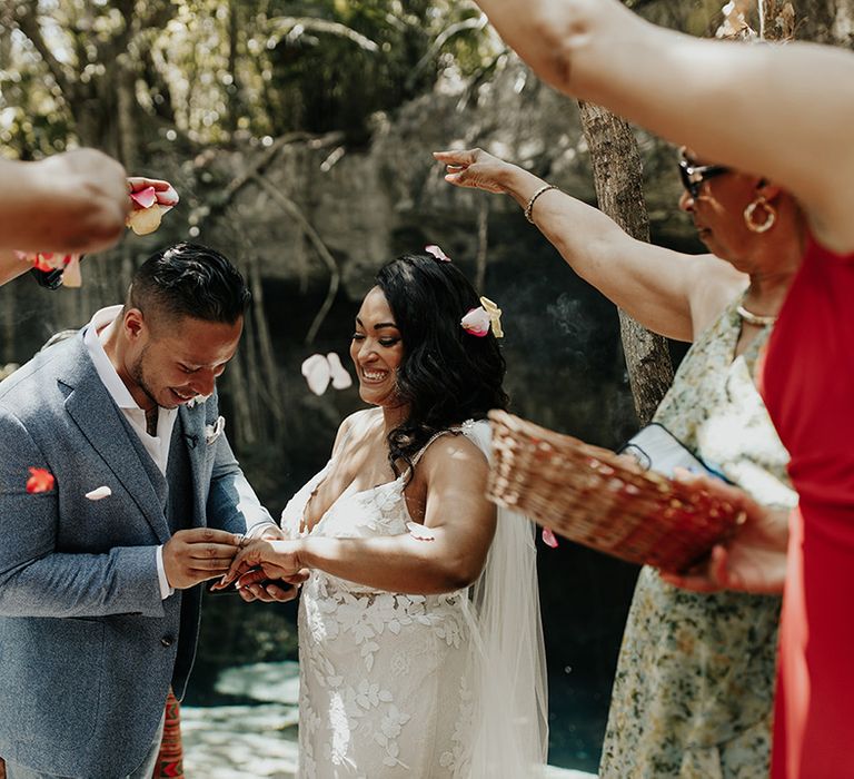 Groom puts on the bride's wedding ring as the guests throw rose petal confetti over the couple